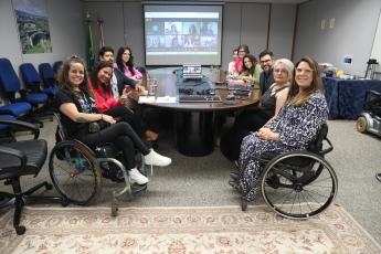 A imagem mostra uma reunião em uma sala de conferências, com cerca de dez pessoas sentadas ao redor de uma mesa oval. Duas mulheres em cadeiras de rodas aparecem em destaque, em primeiro plano, enquanto os demais participantes estão distribuídos ao redor da mesa. Ao fundo, um telão exibe participantes conectados por videoconferência, indicando um encontro híbrido. O ambiente é institucional e transmite uma atmosfera de integração, acessibilidade e colaboração.