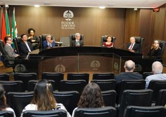 A imagem mostra uma sessão solene em um auditório do Poder Judiciário de Alagoas. Sete pessoas estão sentadas à mesa principal, posicionada em formato semicircular, diante de uma pequena plateia. Ao fundo, destaca-se o brasão e a inscrição “Poder Judiciário de Alagoas” na parede revestida em madeira. O ambiente transmite um caráter institucional, formal e voltado à realização de evento ou cerimônia oficial.