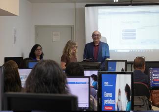 HÁ DUAS MULHERES E UM HOMEM DE PÉ FALANDO PARA VÁRIAS PESSOAS SENTADAS EM UMA SALA DE AULA, COM COMPUTADORES À SUA FRENTE.