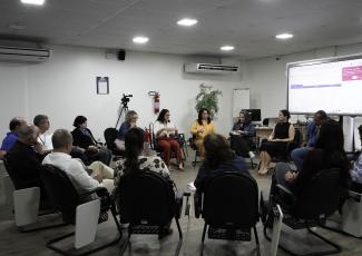 Há várias pessoas sentadas em um círculo. Há uma tela grande para exposição do conteúdo a ser debatido.
