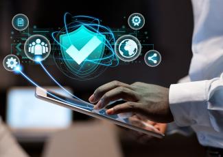 A imagem mostra uma pessoa utilizando um tablet enquanto interage com uma interface digital representada por elementos gráficos em tons de azul. No centro da composição, destaca-se um escudo com um símbolo de verificação, remetendo à segurança e à proteção de dados. Ao redor dele, aparecem ícones que representam documentos, usuários, configurações, certificação, compartilhamento de informações e conexão global, sugerindo conceitos relacionados à segurança cibernética, governança digital.