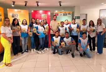 Há um grupo de estudantes e três adultos (um homem e duas mulheres) em uma sala com cartazes