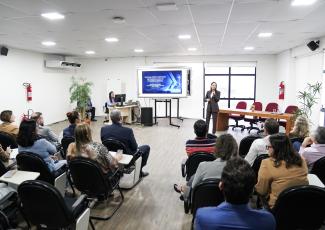 Auditório com várias pessoas sentadas. Elas assitem uma pessoa falar. Há um telão atrás da mulher que fala.