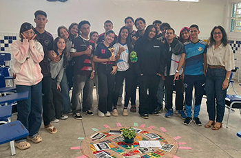 Um grupo de adolescentes e uma professora posam para uma foto em uma sala de aula. À frente deles, no chão, há uma atividade organizada em círculo com cartões coloridos, livros e uma planta ao centro, indicando uma dinâmica ou oficina educativa realizada durante o encontro.