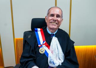 Homem branco, de cabelo grisalho, usando toga preta, sentado e sorrindo