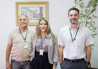 Na fotografia, há três pessoas em pé lado a lado, sorrindo para a câmera, todas utilizando crachás corporativos em cordões escuros no pescoço:  À esquerda: Um homem de cabelos curtos grisalhos, usando uma camisa polo de tom bege/caqui claro.  Ao centro: Uma mulher de cabelos longos, loiros e lisos, vestindo um blazer claro sobre uma blusa estampada.  À direita: Um homem de cabelos curtos, barba e bigode escuros e levemente grisalhos, vestindo uma camisa social de mangas curtas em tom claro (branco ou bege)