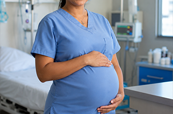 há uma mulher gestante,  com as mãos na barriga, em um ambiente hospitalar