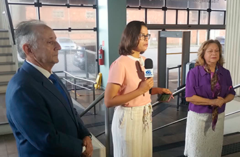 A imagem mostra três pessoas em um ambiente interno. Ao centro, uma mulher segura um microfone durante uma entrevista, enquanto um homem, à esquerda, e outra mulher, à direita, acompanham a gravação. Ao fundo, há grandes portas de vidro.