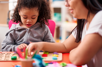 há uma criança (menina) sentada em uma mesinha brincando com jogos, acompanhada de uma mulher