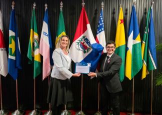 Pessoas Centrais: Uma mulher (à esquerda) vestindo um blazer cinza-claro sobre blusa branca e saia preta, e um homem (à direita) de terno escuro, gravata e óculos de grau. Ambos estão sorrindo e segurando juntos um certificado/diploma do "Ranking da Transparência do Poder Judiciário 2026" (concedido pelo CNJ).  Fundo de Bandeiras: Logo atrás do casal, há um painel ripadinho de tom escuro com vários mastros de bandeiras alinhadas lado a lado, incluindo bandeiras de estados brasileiros