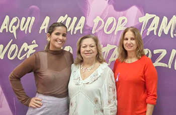 Três mulheres posam para uma foto em frente a um pano de fundo roxo, em plano médio. A mulher da esquerda está sorrindo. Ela tem cabelos castanhos presos e usa um suéter marrom e calças rosa claro. A mulher no centro está sorrindo para a câmera. Ela usa uma blusa com estampa floral e um colar. A mulher da direita está de pé ao lado da mulher central. Ela tem cabelos castanhos na altura dos ombros e usa uma blusa vermelha de mangas compridas e um pingente roxo.