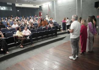 A imagem captura um evento em um auditório, onde um homem de camiseta branca fala ao microfone para uma plateia atenta, tendo ao seu lado três mulheres com vestes coloridas. O público está sentado em poltronas azuis dispostas em fileiras escalonadas, ocupando a maior parte do espaço e voltado para os palestrantes. O ambiente tem paredes de concreto exposto e teto com iluminação embutida e saídas de ar condicionado. No palco, destaca-se o piso de madeira rústica, onde o grupo de oradores está posicionado. 