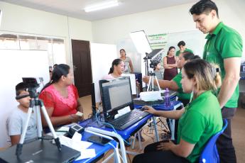 A imagem mostra uma ação de atendimento comunitário em uma sala bem iluminada, onde atendentes de camisa verde operam computadores para cadastrar cidadãos. Em primeiro plano, uma mulher de blusa vermelha e um garoto aguardam enquanto passam por um procedimento com câmera e leitor biométrico. Ao fundo, outra mulher está sentada diante de uma luz de iluminação (softbox) sendo fotografada ou atendida por outra funcionária. Diversas pessoas aguardam de pé e observam o processo, que conta com equipamentos de TI 