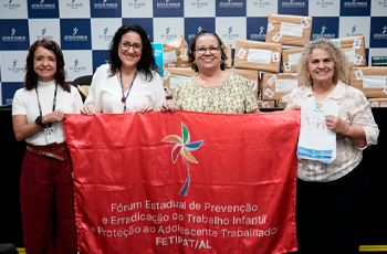 A imagem apresenta quatro mulheres sorridentes alinhadas lado a lado em um ambiente institucional, todas segurando juntas uma grande bandeira vermelha. A bandeira exibe o símbolo de um catavento colorido e o texto do Fórum Estadual de Prevenção e Erradicação do Trabalho Infantil e Proteção ao Adolescente Trabalhador. A mulher da extrema direita também segura alguns papéis ou cartilhas informativas em suas mãos. Ao fundo, observa-se um painel com logotipos da Justiça do Trabalho e do TRT da 19ª Região.