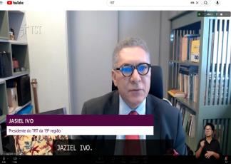 A imagem mostra o presidente do TRT da 19ª Região, desembargador Jasiel Ivo, em uma entrevista por videoconferência. Ele aparece de terno e gravata, diante de uma estante de livros, enquanto uma intérprete de Libras realiza a tradução no canto inferior direito da tela.