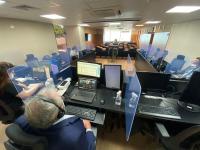 Na sala de sessões do Pleno, um homem de cabelos brancos está sentado numa bancada, olhando para duas telas de computador. Na mesma bancada há outras três pessoas afastadas. As cadeiras do público, ao fundo da imagem, estão vazias.
