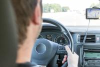 Homem de costas com roupa preta com as mãos no volante dirigindo um carro