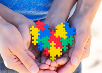 FOTO DO SÍMBOLO DO AUTISMO, COM QUATRO MÃOS SEGURANDO UM CORAÇAÕ COLORIDO