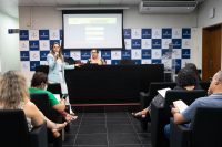 Mulheres e homens sentados, assistindo  à uma mulher de blazer azul, em pé, falar