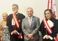 Um homem alto, com cabelo, barba e bigodes brancos usando uma toga com faixa vermelha, uma mulher de vestido preto e branco, morena de cabelos longos e brancos, um homem um pouco mais baixo que o outro, de terno cinza, gravata cinza, blusa branca, cabelos brancos e sem barba e sem bigode e uma mulher loira, de pele branca usando toga e faixa vermelha