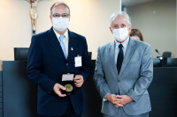 Um homem calvo de terno escuro segurando uma medalha ao lado de um homem mais baixo de cabelos brancos de terno cinza, ambos brancos
