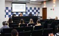 Mulher branca  de roupa lilás no centro da bancada ministrando aula para diversas pessoas 