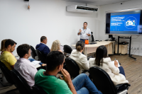 Servidor Rodrigues Rodrigues de camisa cinza claro, à frente explanando o projeto do LIODS por meio de um data show com a projeção de uma tela azul a oito pessoas sentadas.