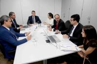 Reunião em volta de uma mesa retangular branca, com 5 homens brancos de terno e duas mulheres brancas