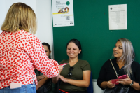 Mulher de blusa vermelha e branca entregando filpeta para três mulheres sentadas trajando roupas escuras.