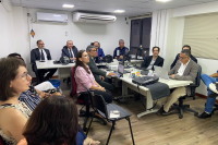 Vários homes de ternos escuros e um de palitó claro, além de duas mulheres, sendo uma com blusa rosa e outra de blusa estampada. Todas as pessoas estão assitindo uma apresentação com informações