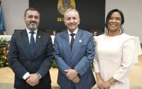 Dois homens de ternos, sendo um vestido na cor escura e outro na cor mais clara. Há uma mulher ao lado com vestido claro. 
