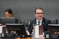 Duas pessoas de pele clara em frente aos monitores de computador, sendo uma mulher com cabelos escuros longos e um homem de camisa branca e terno escuro.