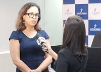Mulher de pele morena, de cabelos longos e pretos, usando óculos de armação preta e um vestido azul marinho concedendo entrevista a uma repórter de cabelos pretos.