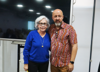 Mulher de cabelos brancos, blusa de mangas longas azul e óculos de armação preta ao lado de homem calvo, barbudo de camisa estampada. 