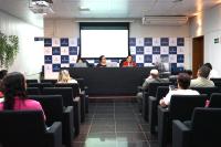 Mesa de auditório composta com três pessoas  de pele clara, sendo duas delas de roupa escura e uma de blusa vermelhas falando para divresas pessoas na plateia