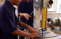 Duas pessoas com peles claras vestidas de roupas na cor azul manuseando uma máquina de cor bege e amarela