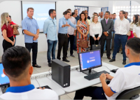 Homens e mulheres de várias etnias de pé trajando roupas em tons claros e escuros ouvindo homem falando, de frente a dois homens jovens sentados a frente de computadores, vestidos de camisas nas cores branca e azul.