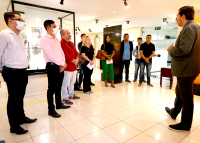 Homens e mulheres de pé,  vestidos com roupas de diversas cores, assistindo apresentação de homem branco de terno escuro