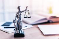 Escultura da Justiça sobre a mesa. Ao fundo, estão desfocados um livro aberto, papeis de anotação e uma caneta.