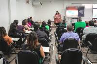 Auditório com muitas pessoas e ao fundo uma palestrante com blusa colorida.
