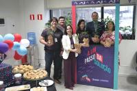 Homens e mulheres posando para foto com mudas de plantas nas mãos. Há balões coloridos e uma mesa com pães e salgados.