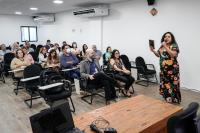 Mulher negra de cabelos cacheados pretos falando na frente de um auditório para várias pessoas sentadas