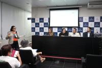 Mulher branca de óculos e cabelos pretos apresentando um slide, quatro pessoas em uma bancada e um auditório cheio de pessoas