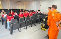 Auditório com várias pessoas sentadas, vestidas com blusas vermelhas e dois homens, à direita, em pé, com vestes na cor laranja.