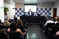 Homem de terno azul escuro e camisa branca falando para público sentado em cadeiras pretas em um auditório.