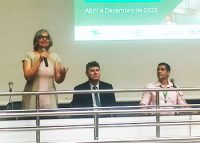Mulher branca de vestido verde e rosa de pé falando. Há dois homens sentados, sendo um de terno escurp e outra de camisa rosa claro