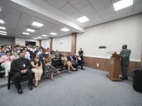 Foto em diagonal registrando várias mulheres e homens sentados em um auditório. Na frente das pessoas, em um púlpito, está o presidente do TRT-11, desembargador Audaliphal Hildebrando, um homem negro e de terno cinza. 