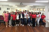 Foto com muitas pessoas, homens e mulheres, em pé. Piso de madeira e parede, ao fundo, na cor branca.