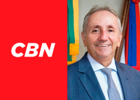 Foto de um homem branco de cabelo branco e terno azul ao lado de um card vermelho com as letras CBN em branco