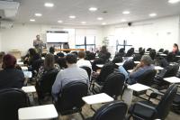 Foto mostra auditório com muitas cadeiras e algumas pessoas sentadas. No alto da foto, um palestrante em pé.